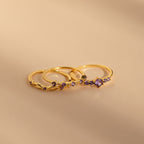 The Amethyst Stacking Ring Set features three delicate gold rings with amethyst stones, elegantly displayed on a smooth beige surface—ideal for lovers of fashion-forward jewelry.