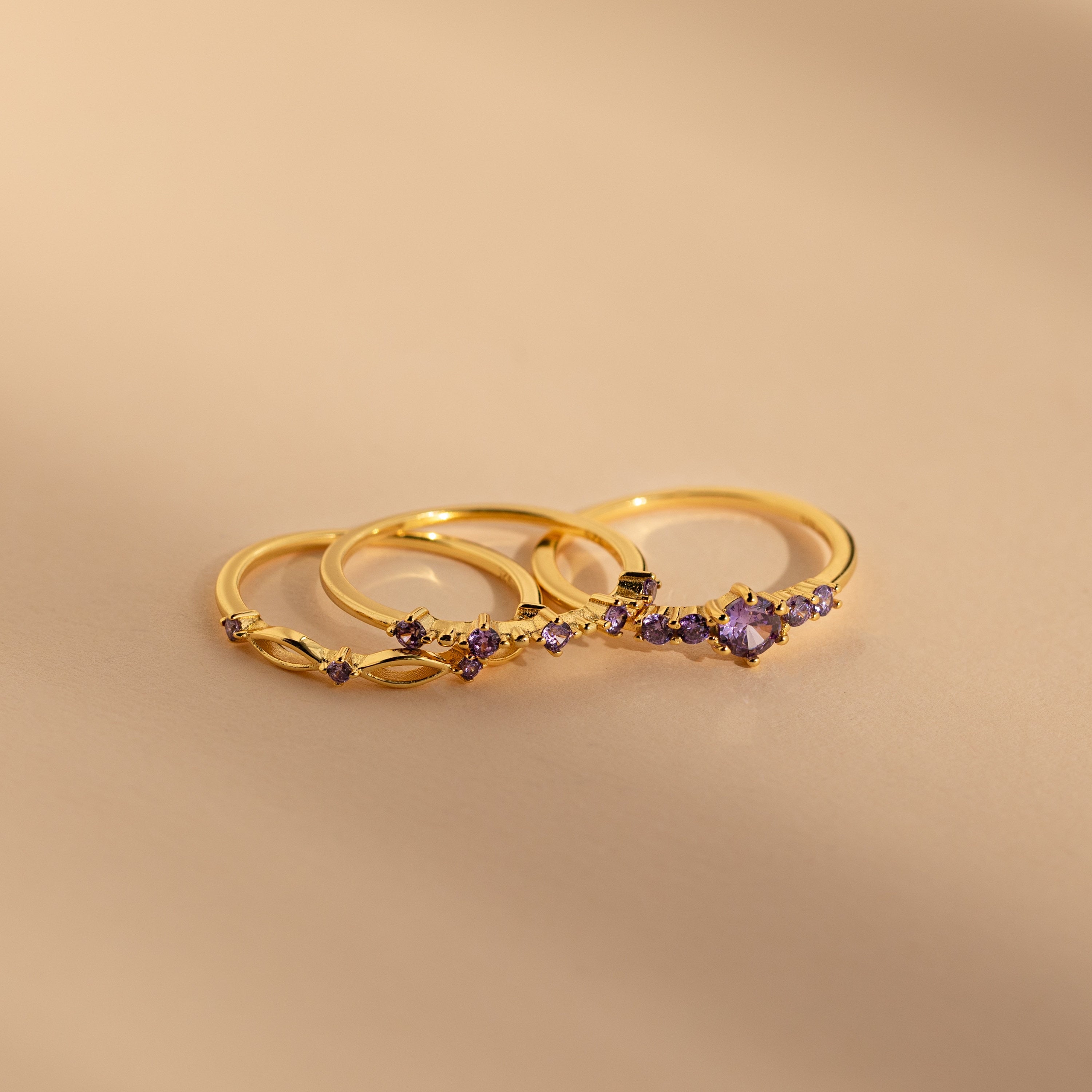 The Amethyst Stacking Ring Set features three delicate gold rings with amethyst stones, elegantly displayed on a smooth beige surface—ideal for lovers of fashion-forward jewelry.