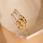 The Emerald Stacking Ring Set features three gold rings with green and white gemstones, displayed on a light textured surface. Ideal for lovers of Art Deco jewelry and vintage-inspired elegance.