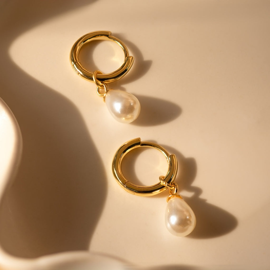 Dainty Pearl Drop Huggies: Elegant gold hoop earrings featuring teardrop pearls, shown on a beige surface with soft shadows—ideal for adding a refined touch to any occasion.