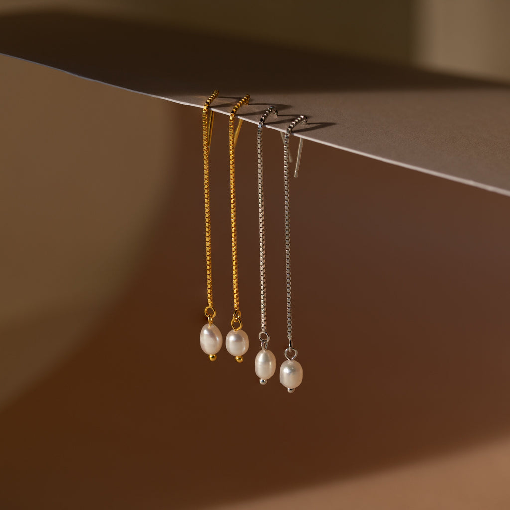 Two pairs of Pearl Threader Earrings—one in gold, one in silver—are elegantly displayed, hanging from the edge of a surface.
