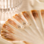 Dainty Knot Pearl Earrings in Sterling Silver displayed on a brown and cream seashell with a glass in the background, highlighting their classic charm and timeless sophistication.