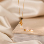 The Pearl Knot Necklace & Earrings Set in 18K Gold, displayed on a beige textured book, makes an elegant gift for your wife—perfect for any special occasion.