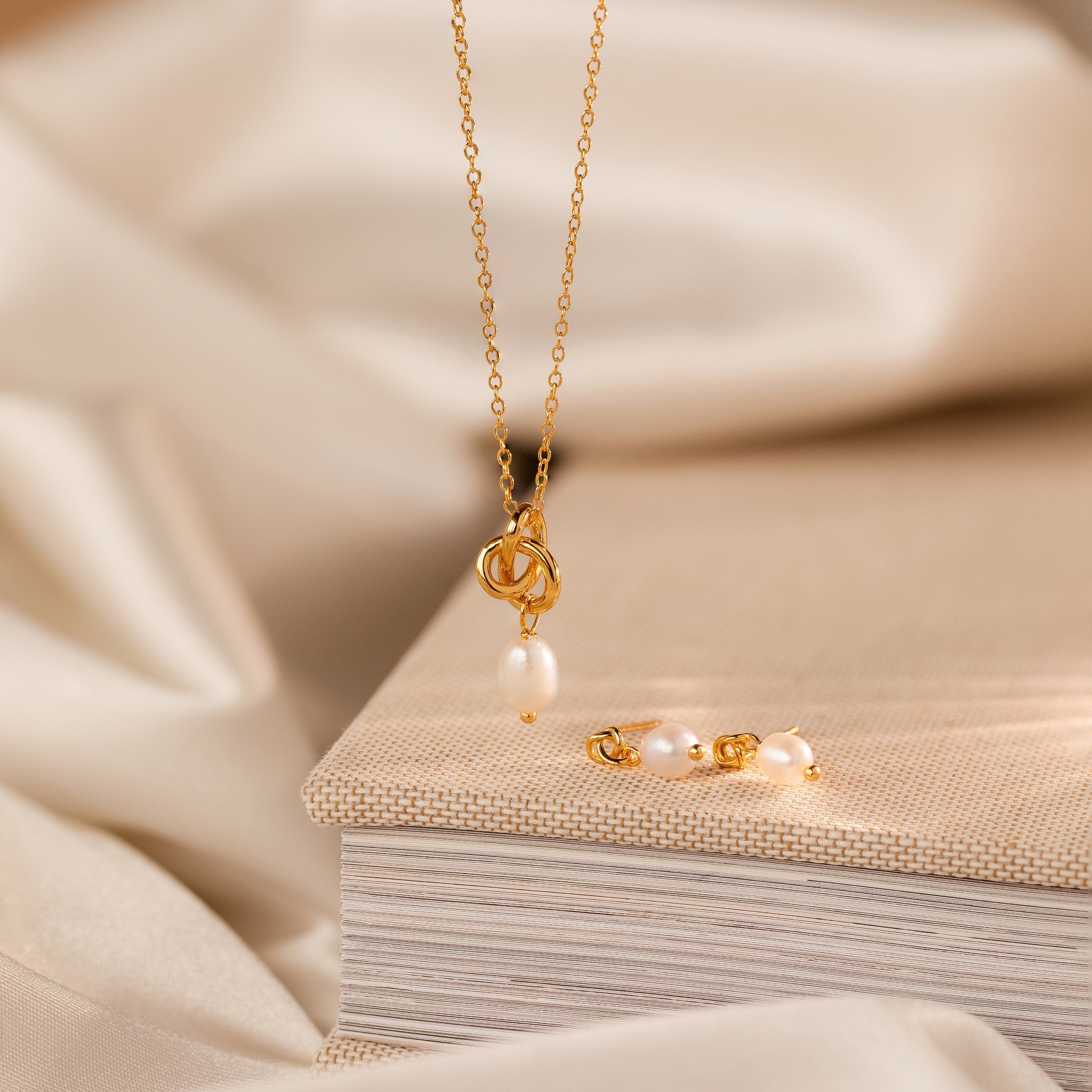The Pearl Knot Necklace & Earrings Set in 18K Gold, displayed on a beige textured book, makes an elegant gift for your wife—perfect for any special occasion.