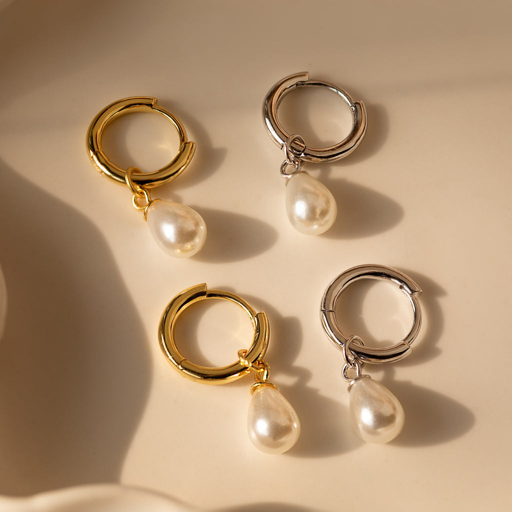 The Dainty Pearl Drop Huggies, featuring dangling teardrop pearls in two gold and two silver styles, are elegantly displayed on a beige surface.