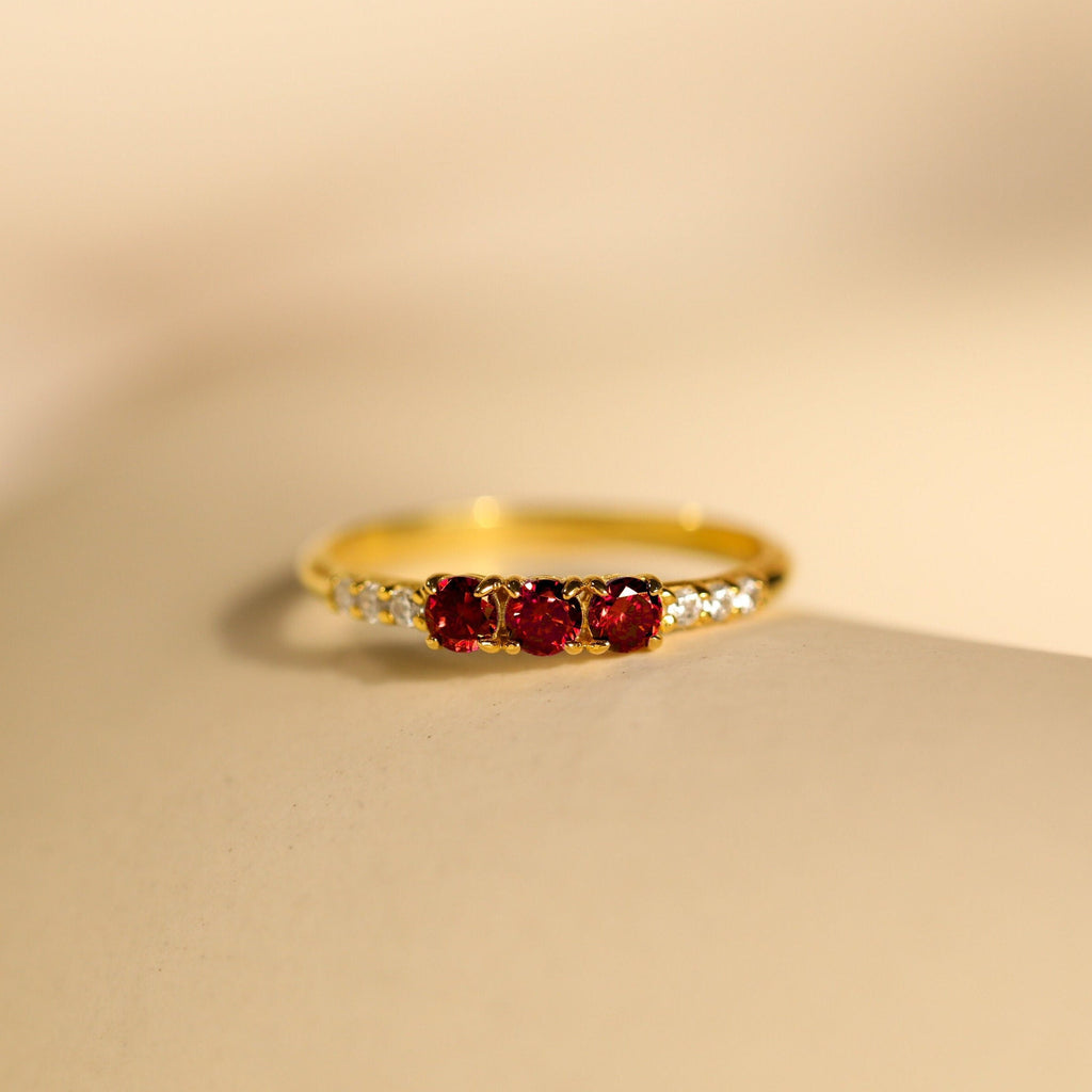 The Pave Triple Birthstone Ring features three red gemstones and small clear stones on the band, shown on a beige surface—ideal for personalized jewelry lovers seeking a customizable birthstone ring.