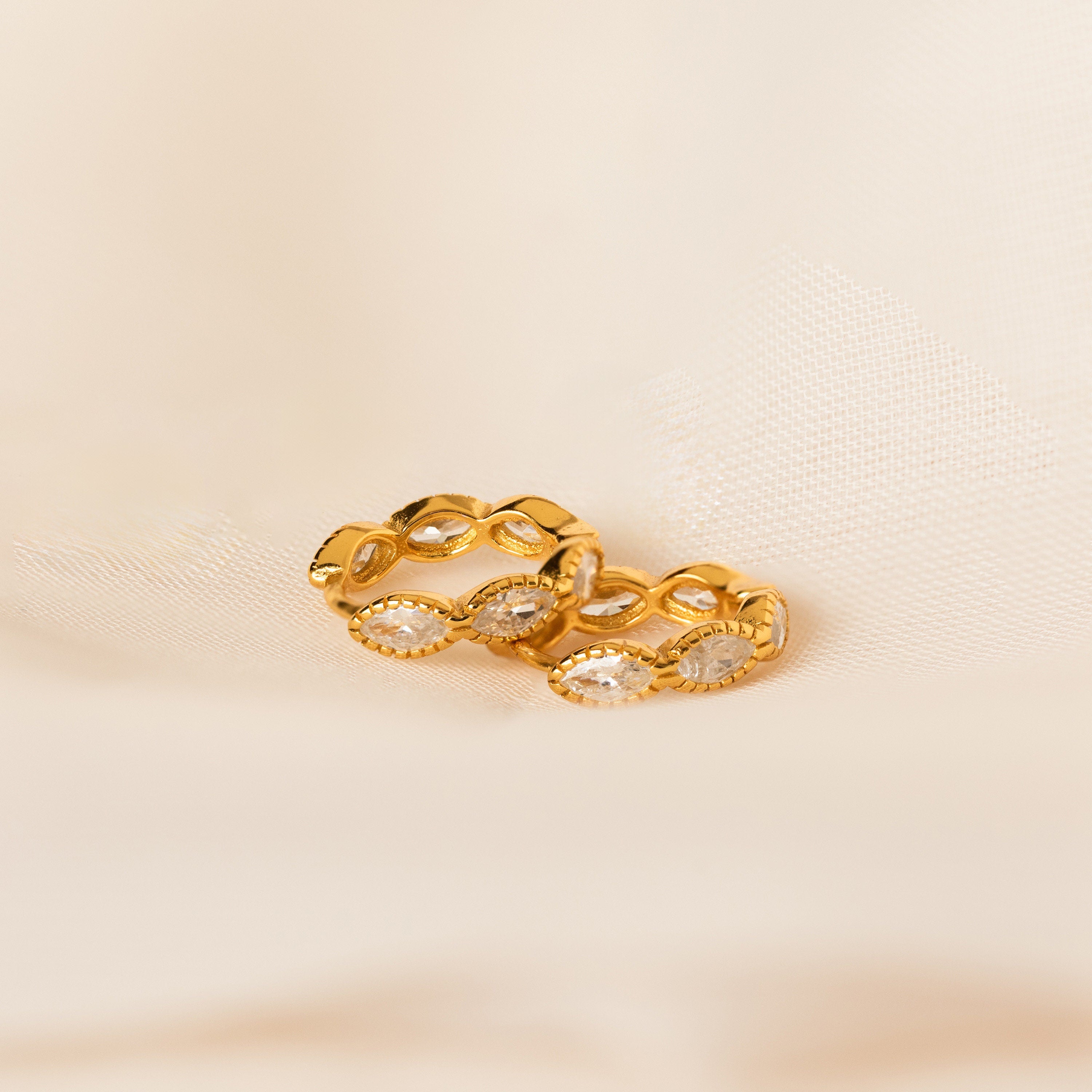 A pair of Marquise Inlay Huggies with sparkling clear stones is displayed on a soft, cream-colored fabric background.