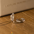 The Marquise Inlay Huggies, two silver hoops with intricate designs, are displayed on a wooden surface bathed in warm light.