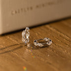 The Marquise Inlay Huggies, two silver hoops with intricate designs, are displayed on a wooden surface bathed in warm light.