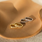 Two pairs of Baguette Link Hoops—one gold, one silver—adorned with embedded stones rest on a tan ceramic dish. Their chic, minimalist design makes them perfect for any occasion.