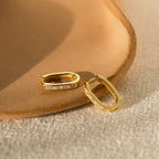 The Baguette Link Hoops, featuring gold with embedded stones and a geometric minimalist design, are displayed on a textured beige surface.