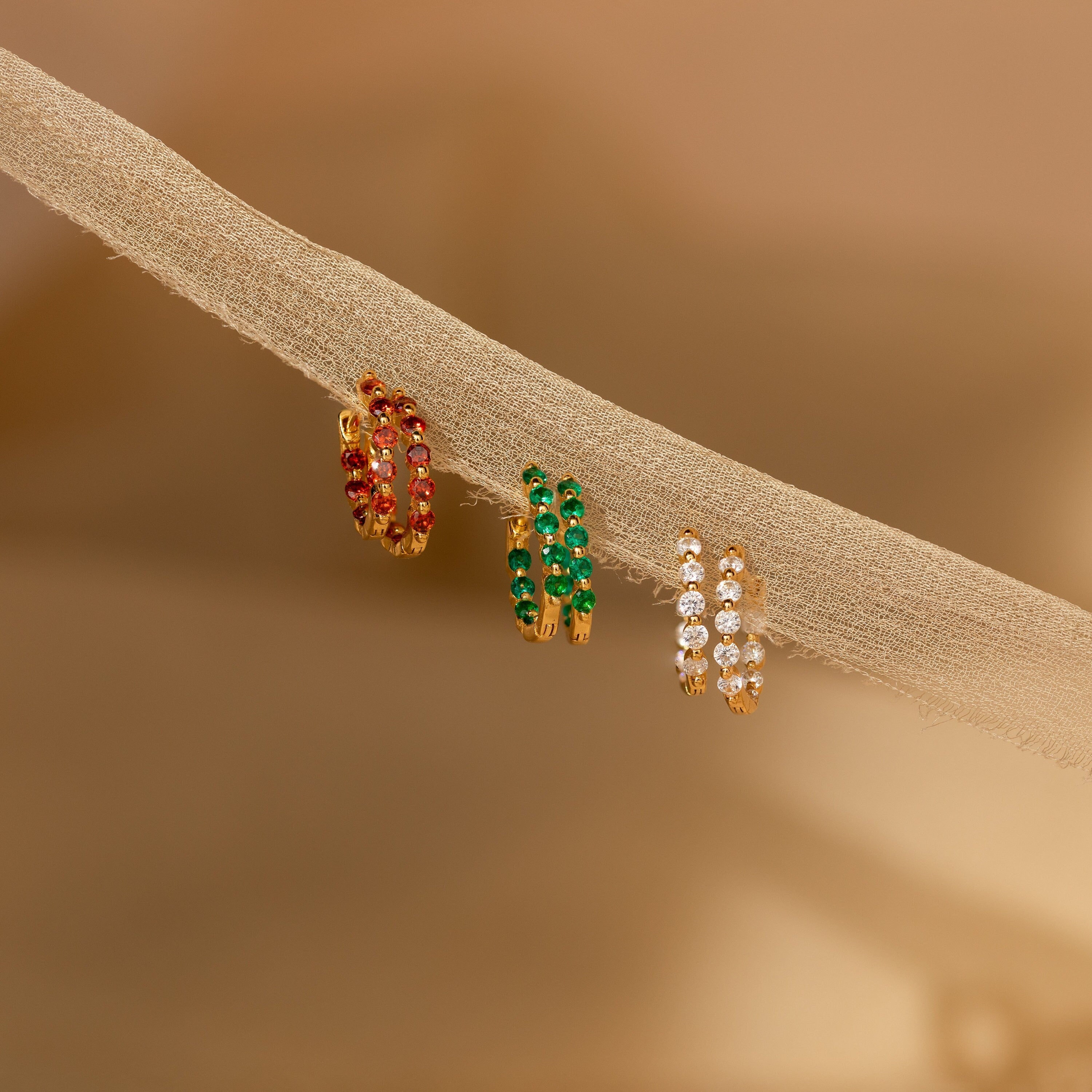 The Inside Out Garnet Huggies showcase three gold hoop earrings adorned with red, green, and clear stones, adding vibrant color to the design and beautifully displayed on a beige fabric branch.