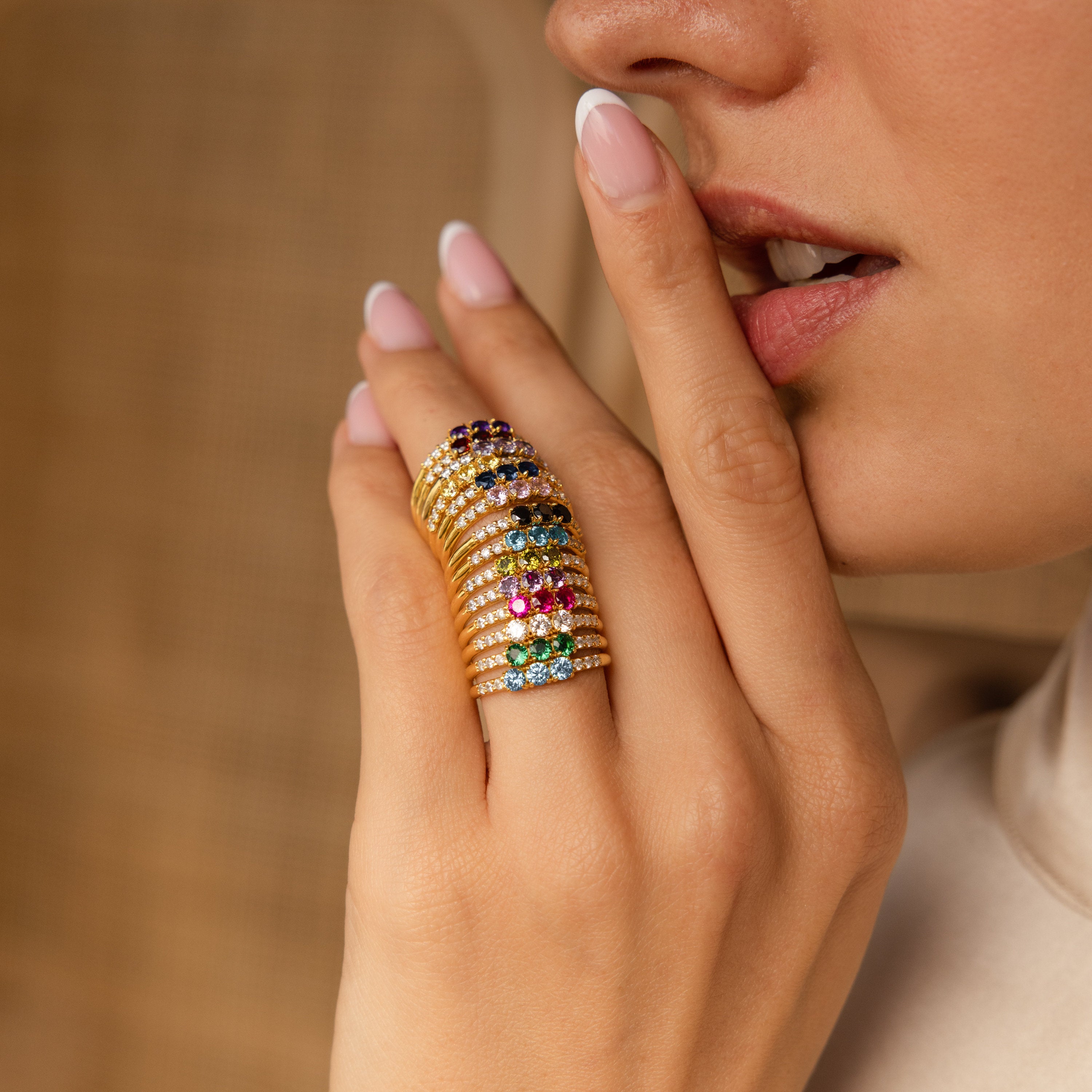 Woman with manicured nails wears multiple colorful gemstone rings, including the Pave Triple Birthstone Ring, as she gently touches her lips.