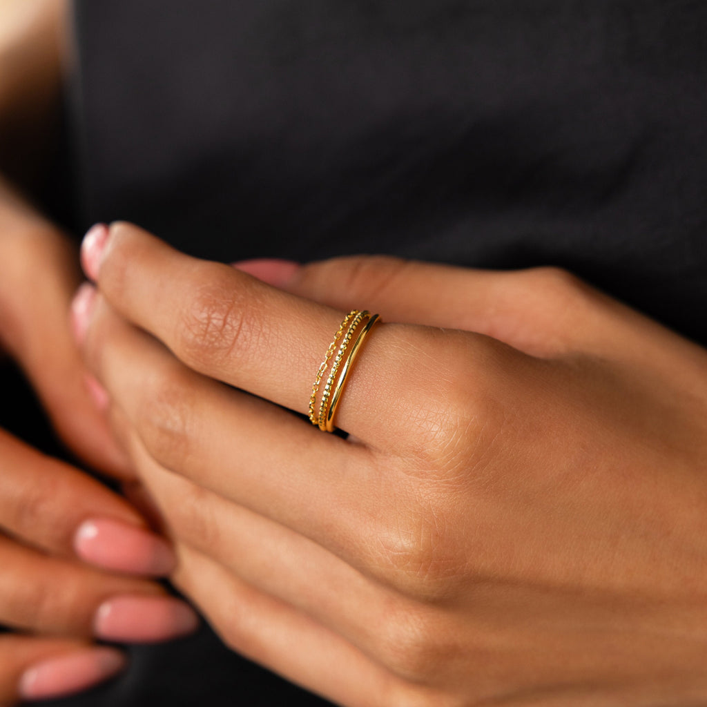 A hand with pink nails displays the Textured Stacking Ring Set—delicate gold bands with subtle texture—against a dark background, ideal for those who appreciate minimalist style.