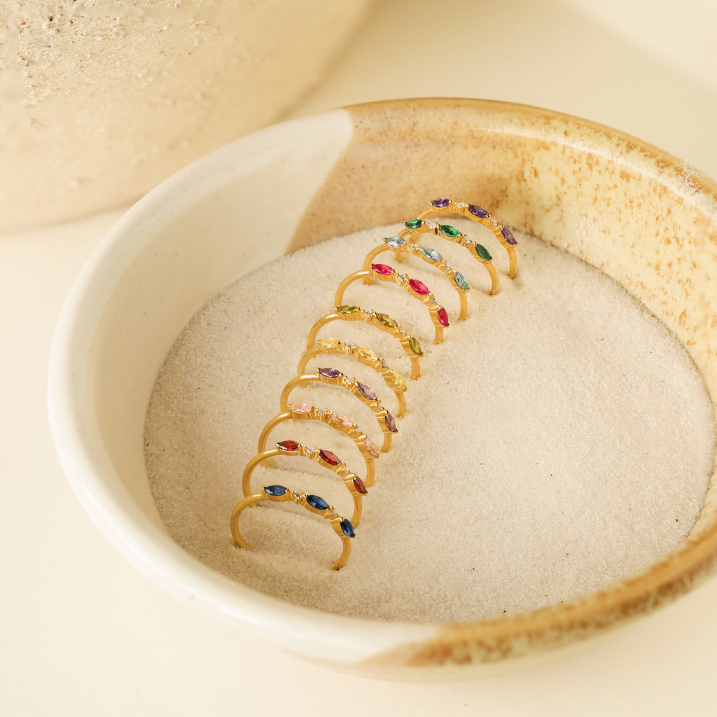 A Triple Marquise Birthstone Ring with colorful CZ Diamond gemstones is displayed in a ceramic bowl filled with sand.