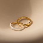Two wavy rings, one gold and one white, overlap on a beige surface to highlight the Duo Wave Ring Set—a stylish choice for fans of minimalist jewelry.