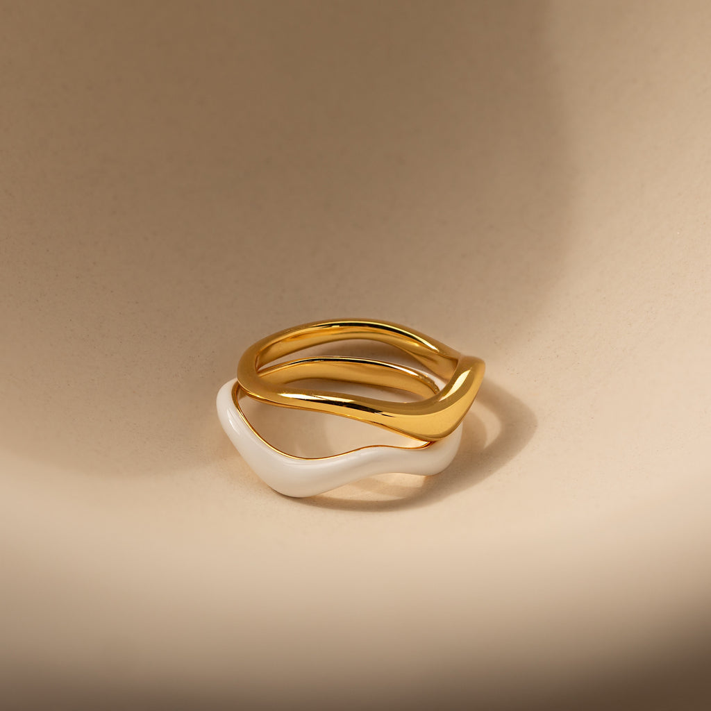 The Duo Wave Ring Set features two wavy rings—one gold, one white—stacked elegantly on a beige surface, perfect for fans of minimalist jewelry.