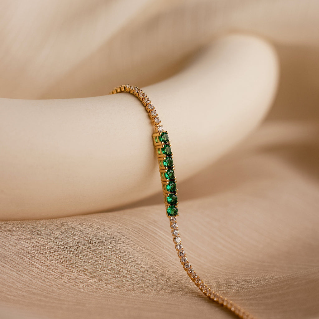 The Emerald Diamond Tennis Bracelet features sparkling diamonds and vibrant emeralds, elegantly showcased on a smooth, beige, curved surface.