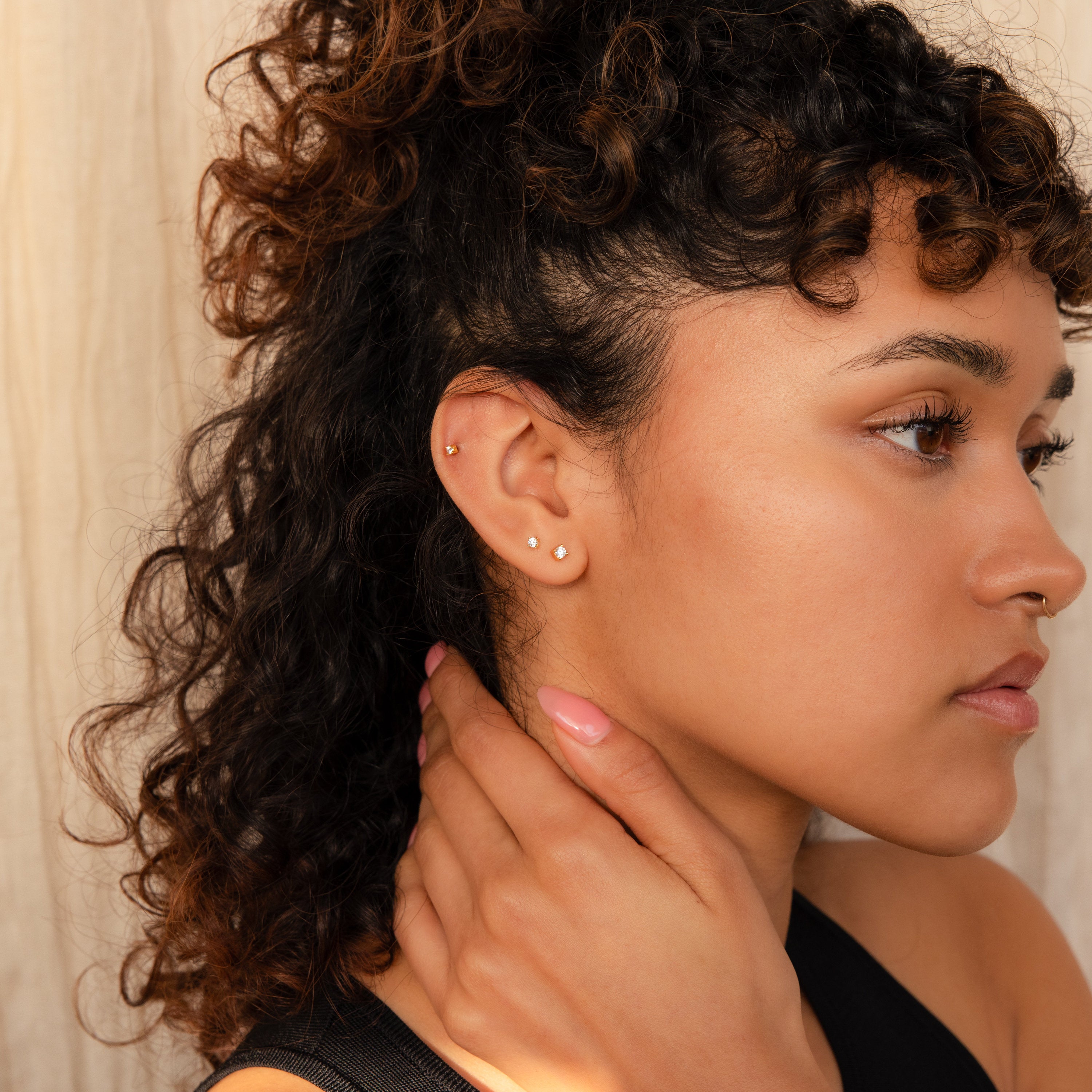 A woman with curly hair and multiple piercings touches her neck, showcasing dainty earrings and the Diamond Flatback Studs Set as she gazes to the side.