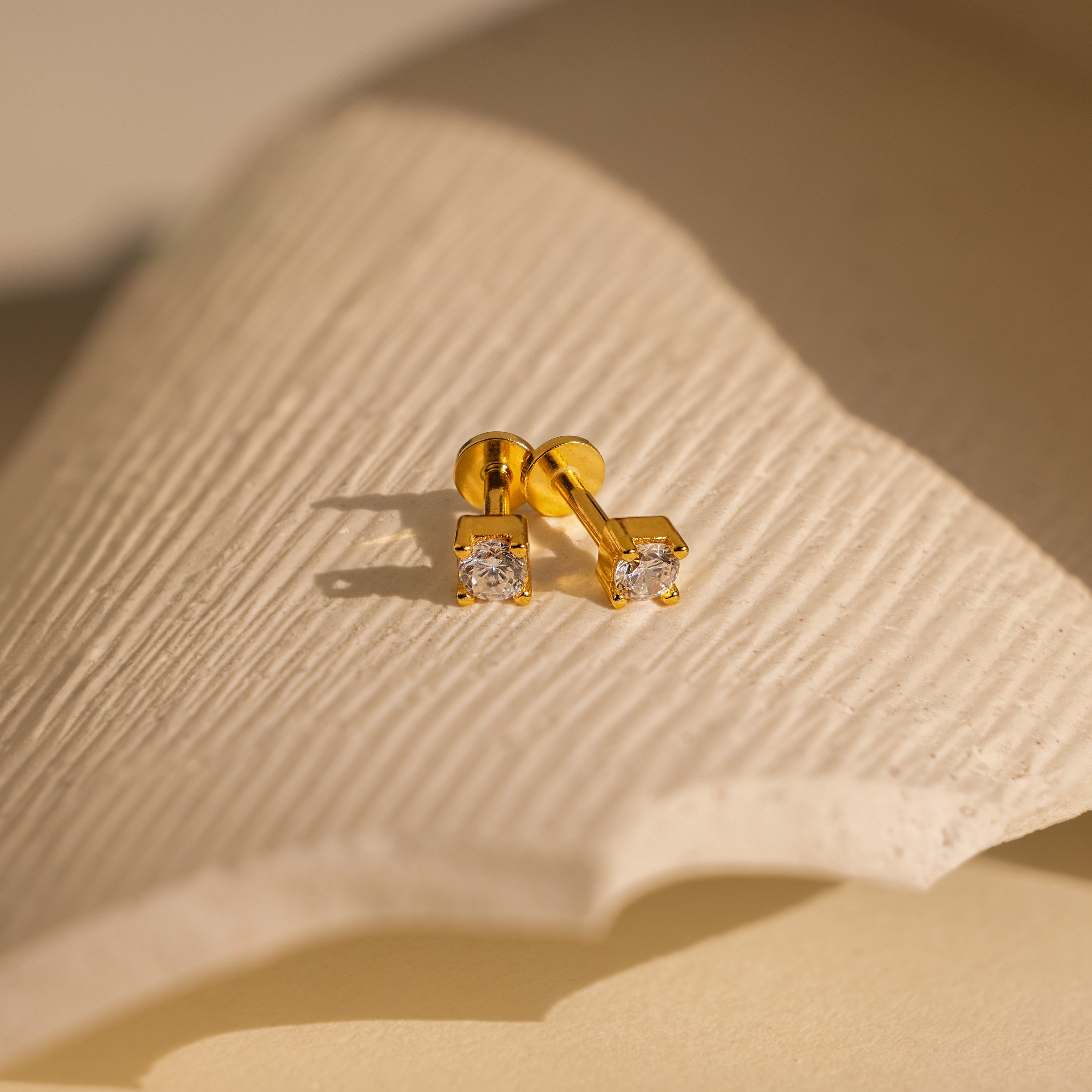 A pair of Diamond Flatback Studs set in 18K gold with secure screw back design, featuring clear gemstones and displayed on a textured, cream-colored surface.