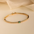 The Emerald Baguette Tennis Bracelet features small round CZ diamond stones and a single rectangular green emerald, displayed on beige fabric—ideal for statement jewelry lovers.