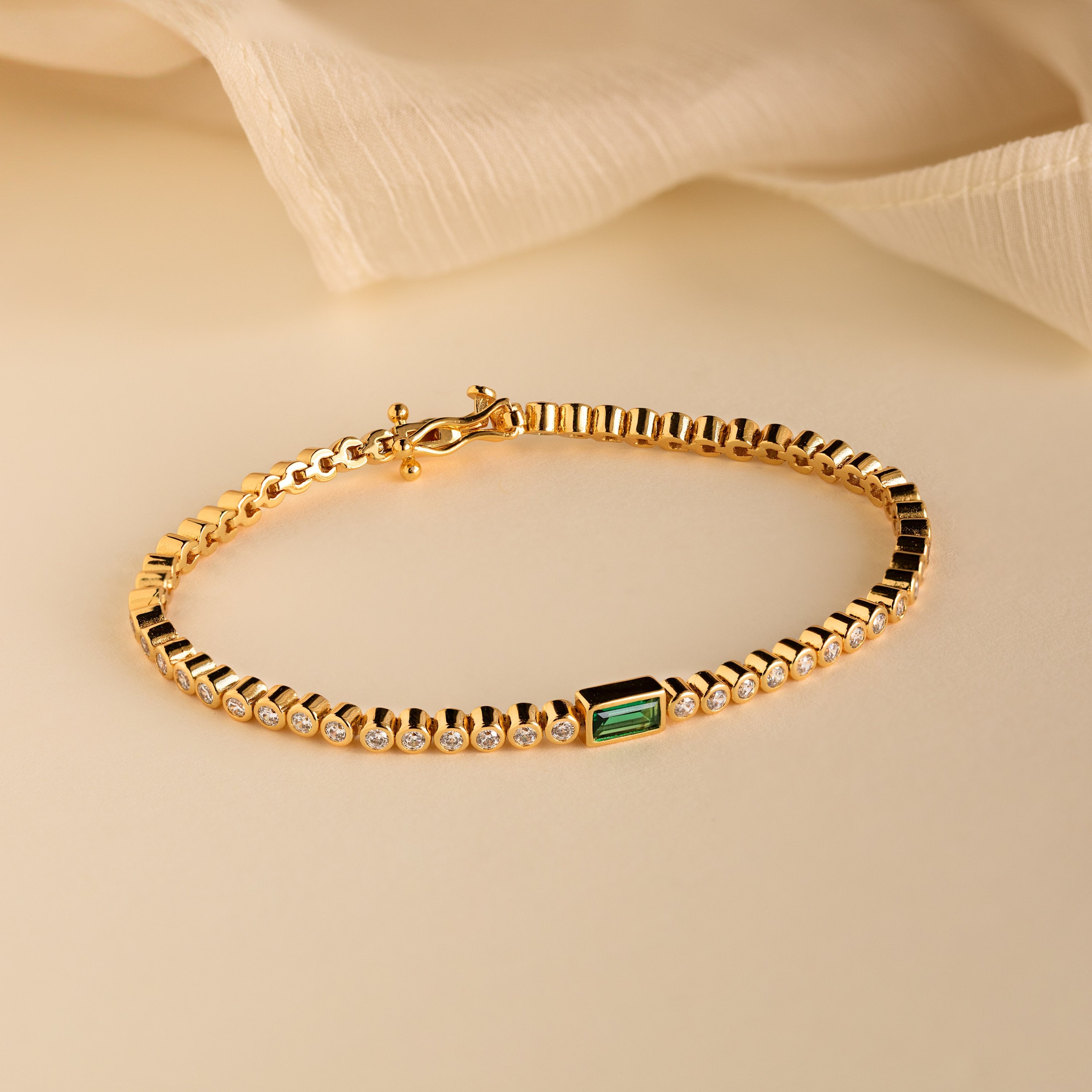 The Emerald Baguette Tennis Bracelet features small round CZ diamond stones and a single rectangular green emerald, displayed on beige fabric—ideal for statement jewelry lovers.