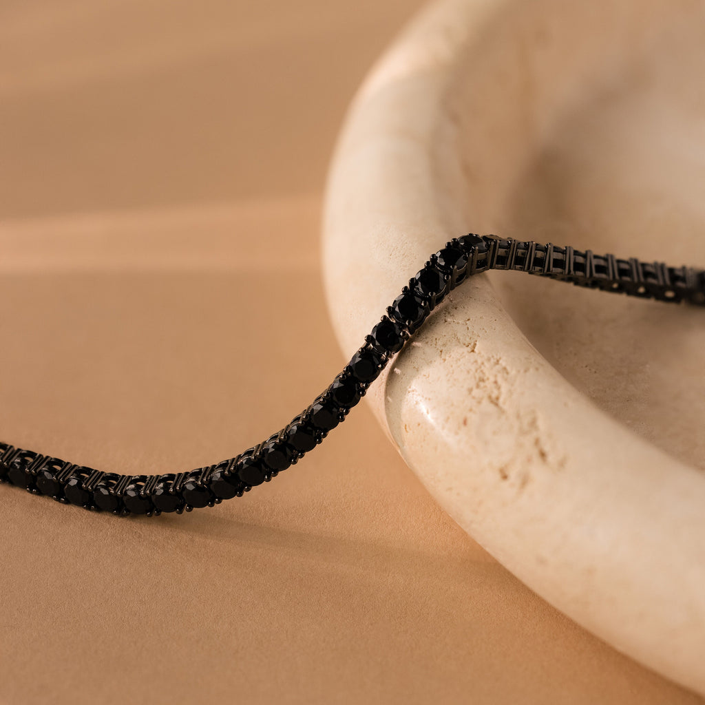 The Onyx Tennis Bracelet sparkles with Black CZ Diamonds, displayed elegantly atop a smooth, round, cream-colored marble dish.