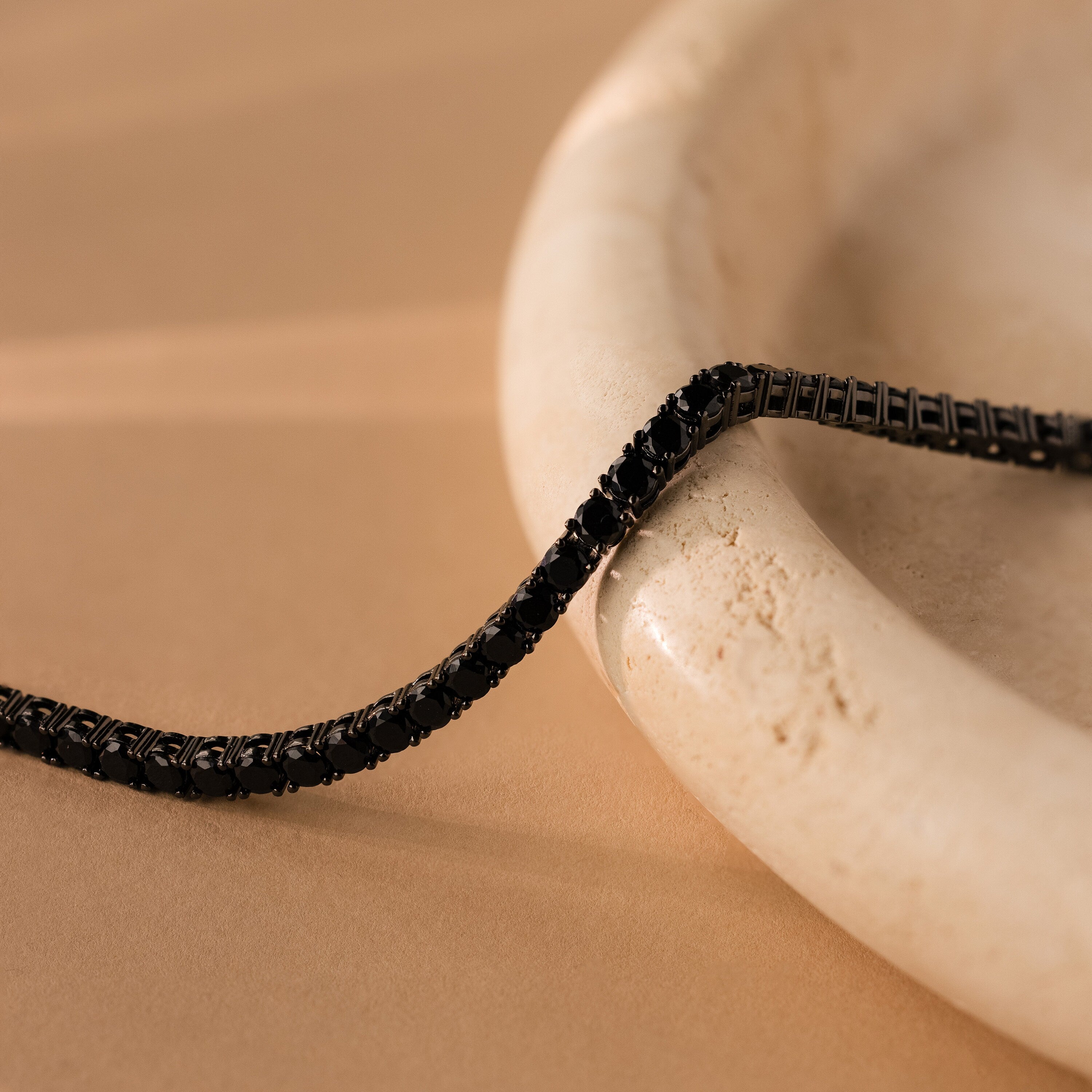 The Onyx Tennis Bracelet sparkles with Black CZ Diamonds, displayed elegantly atop a smooth, round, cream-colored marble dish.