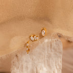 The Marquise Diamond Earrings Set, ideal as bridesmaid gifts, features gold hoops with clear gemstones and is displayed on a textured stone and beige fabric background.