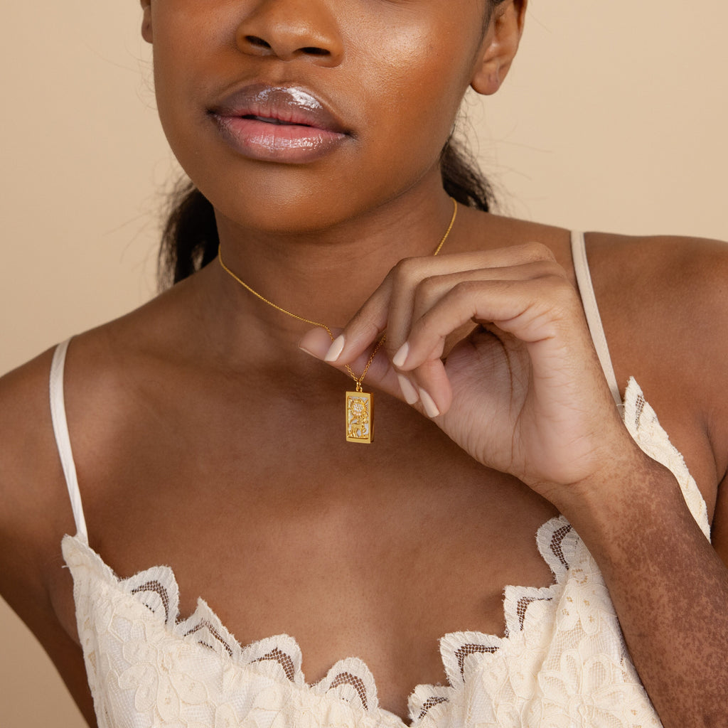 Woman in a cream lace top holding the Sun & Moon Tarot Necklace—a rectangular gold pendant near her chest, making a stunning statement.