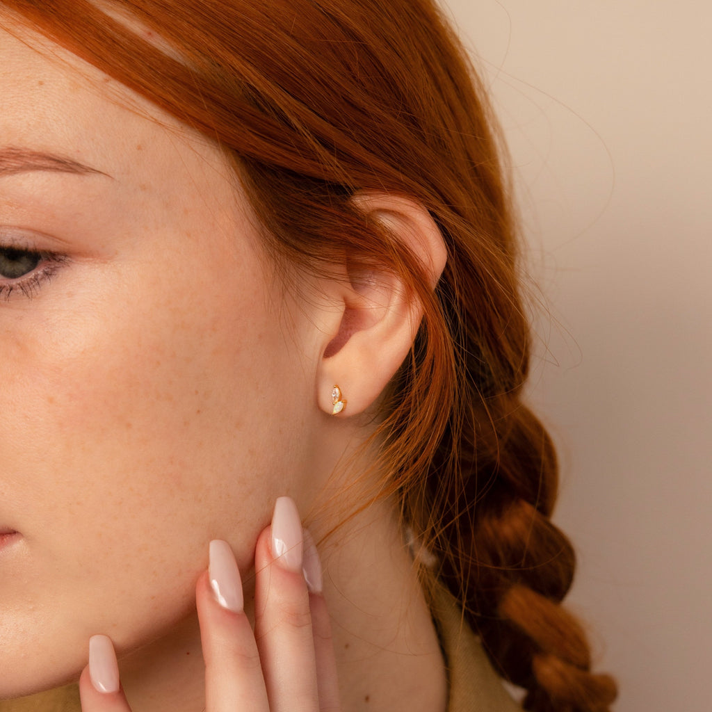 A woman with red hair in a braid touches her face while wearing the elegant Opal & Diamond Marquise Studs—an ideal and unique bridesmaid gift.