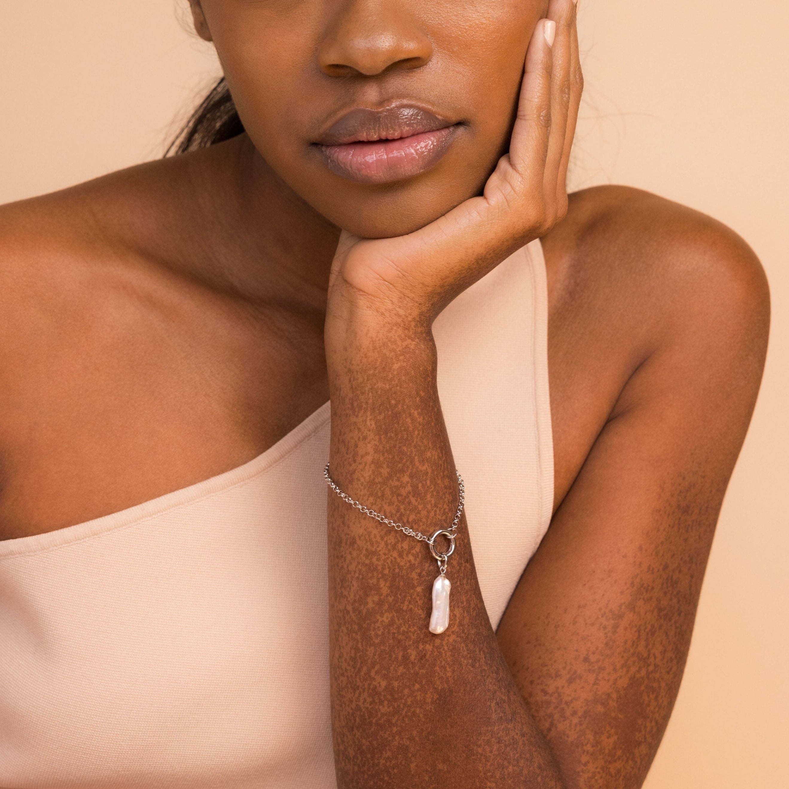 Woman in a one-shoulder beige top rests her face on her hand, wearing a silver bracelet featuring the Long Baroque Pearl Charm.