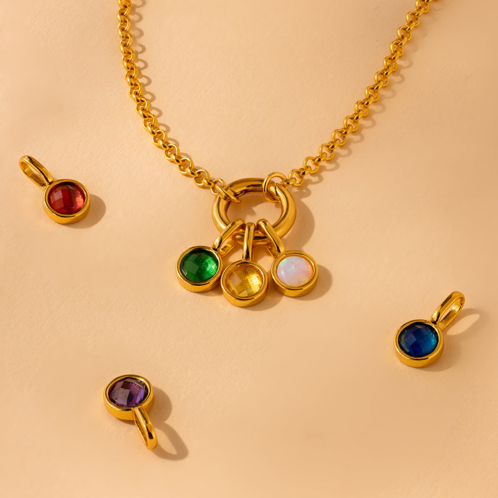 Emerald, Topaz and Opal signet birthstone charms attached to a gold rolo charm necklace. Other signet birthstone charms arranged next to the necklace