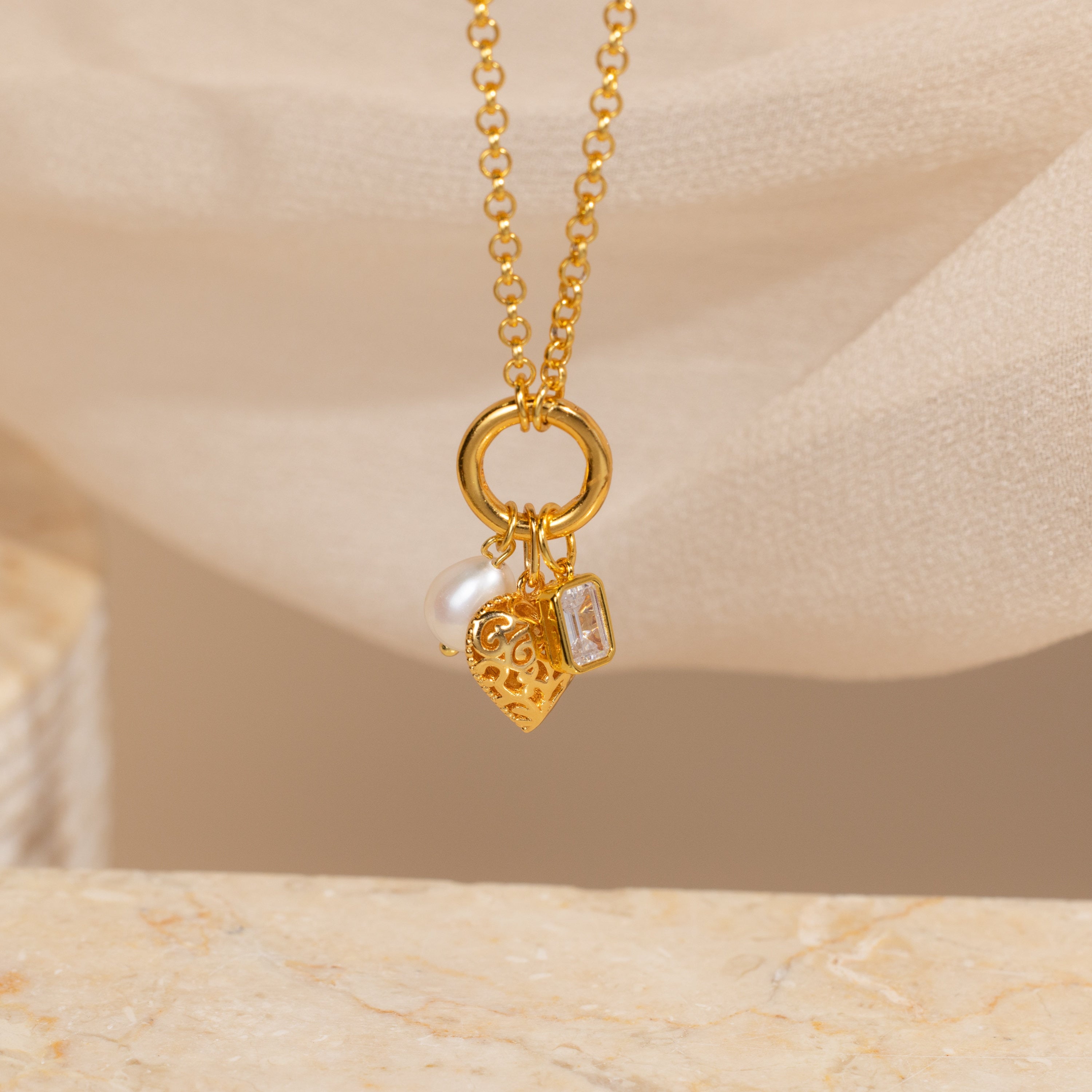 The Rolo Circle Charm Necklace features a gold Rolo Link Chain with a heart charm, pearl, and rectangular gem pendant, elegantly displayed on soft beige fabric.