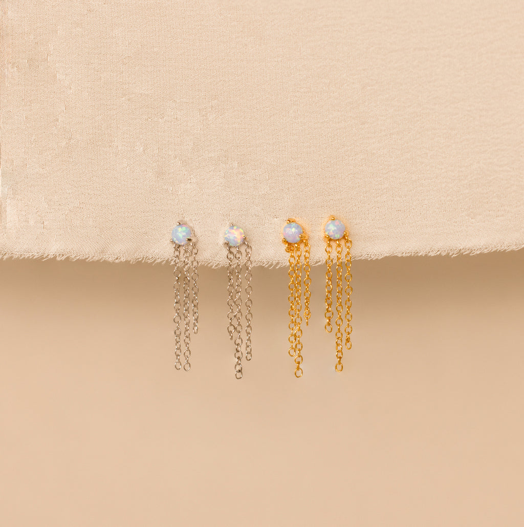 Opal Chain Earrings in both silver and gold are displayed on beige fabric, featuring stud opals with dangling chains—a beautiful addition to any jewelry collection.