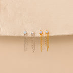 Opal Chain Earrings in both silver and gold are displayed on beige fabric, featuring stud opals with dangling chains—a beautiful addition to any jewelry collection.