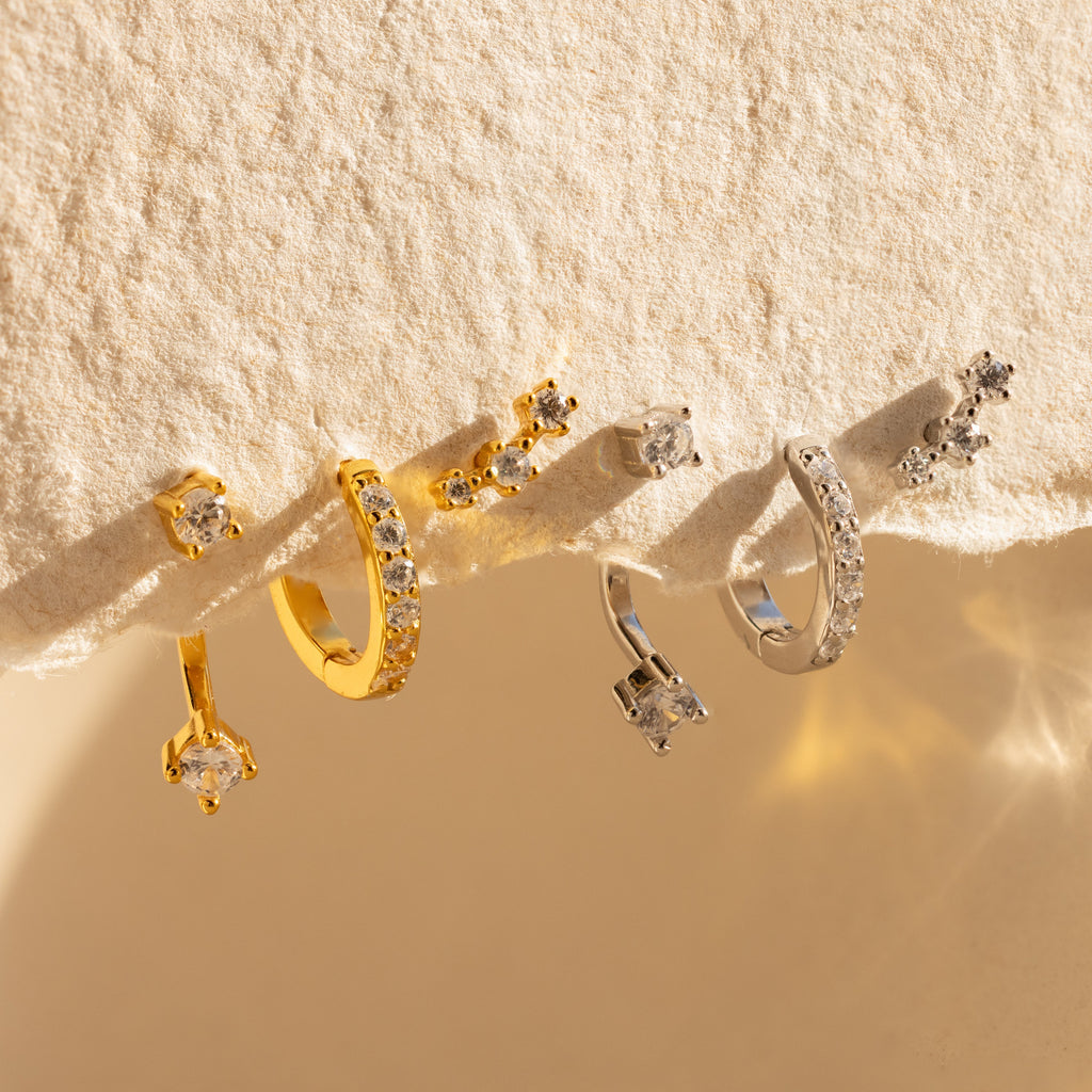 Two pairs of Diamond Earrings Set in 18K Gold, featuring sparkling pave hoops, are displayed on a textured cream surface.