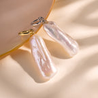Two Long Baroque Pearl Charms with gold and silver loops rest on a pale, reflective surface, highlighting the unique allure and timeless appeal of these distinctive pearl jewelry pieces.