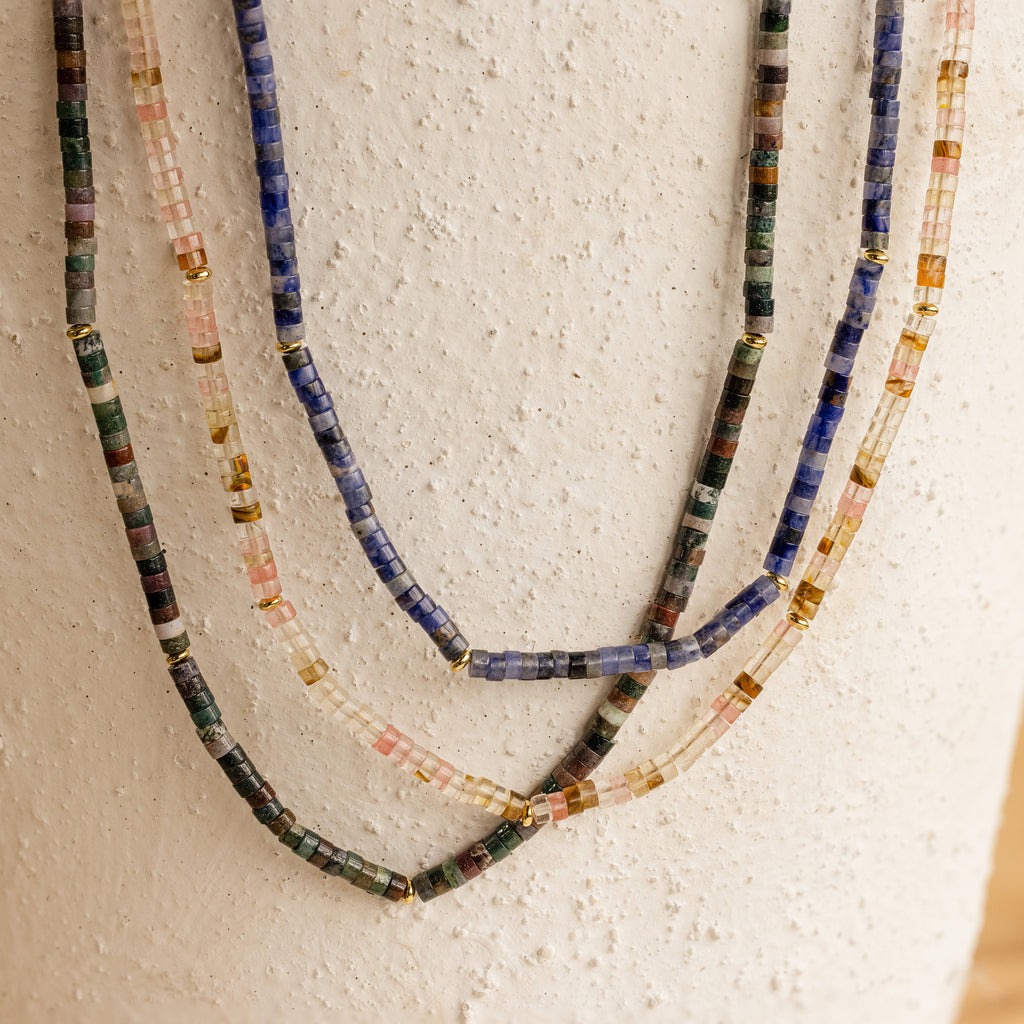 The Rose Quartz Beaded Necklace, featuring colorful beads and gold accents, hangs against a textured white surface—an elegant gift for a best friend.