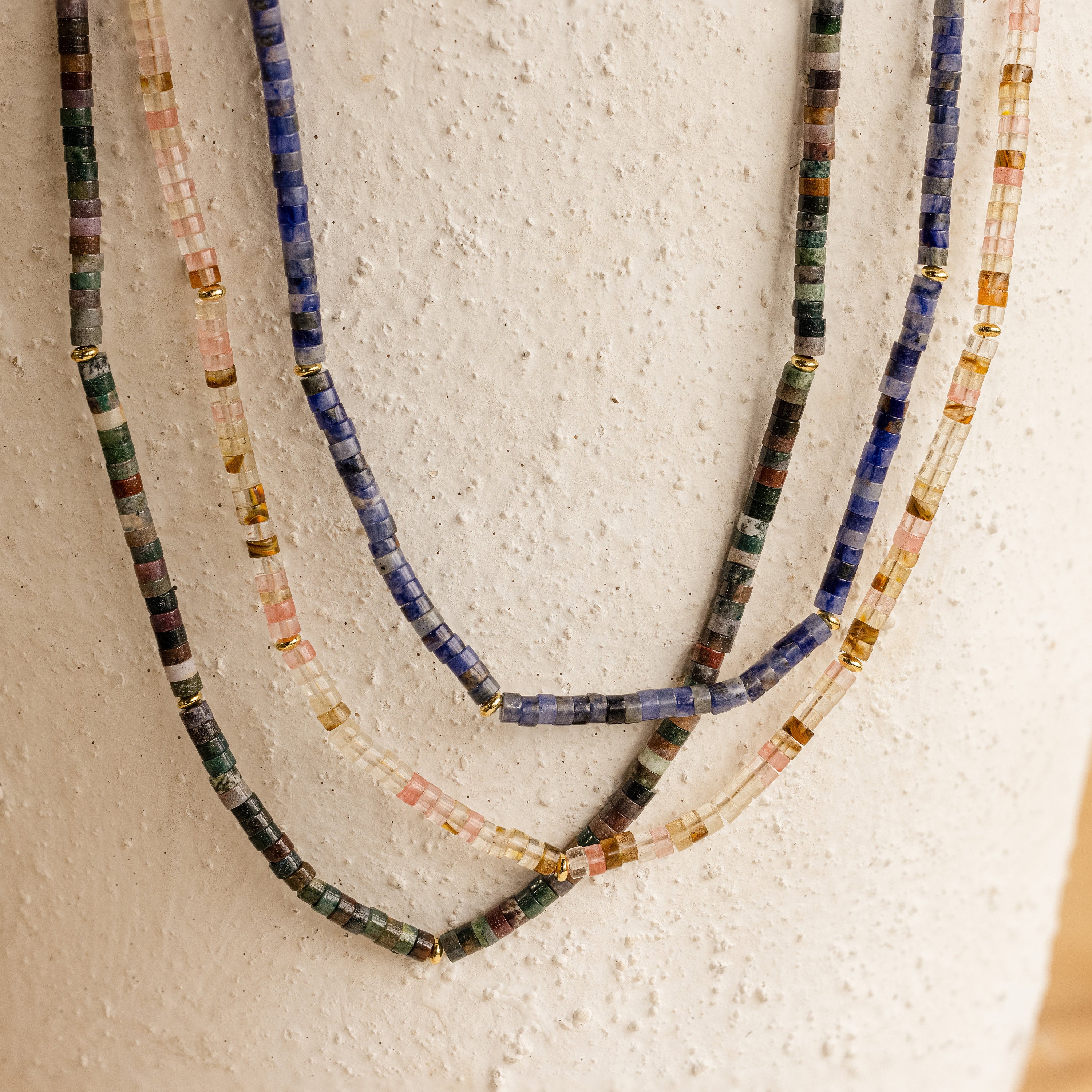 The Rose Quartz Beaded Necklace, featuring colorful beads and gold accents, hangs against a textured white surface—an elegant gift for a best friend.