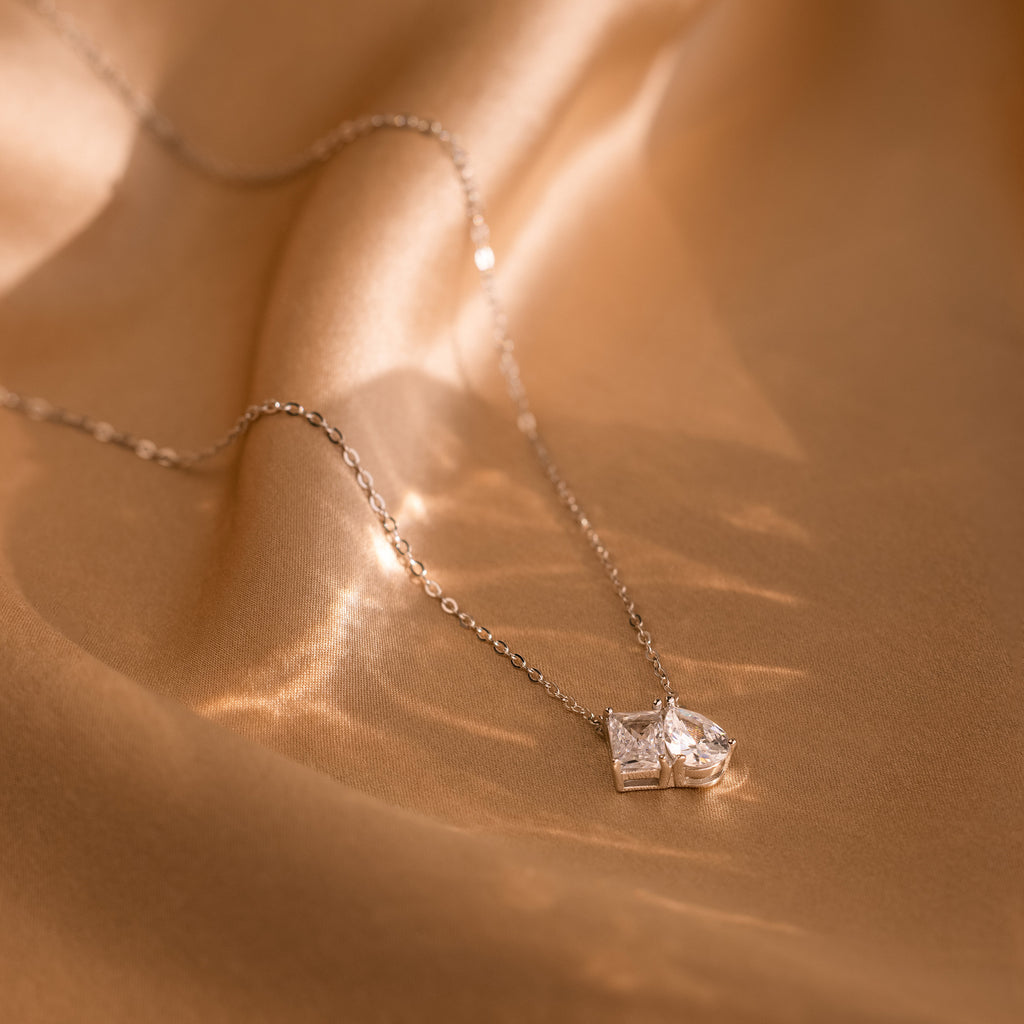 The Toi et Moi Diamond Necklace, featuring two square gemstones, is displayed on smooth, golden fabric.