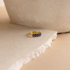 The Oval Birthstone Ring, adorned with five blue gemstones, is displayed on a textured, off-white stone surface.