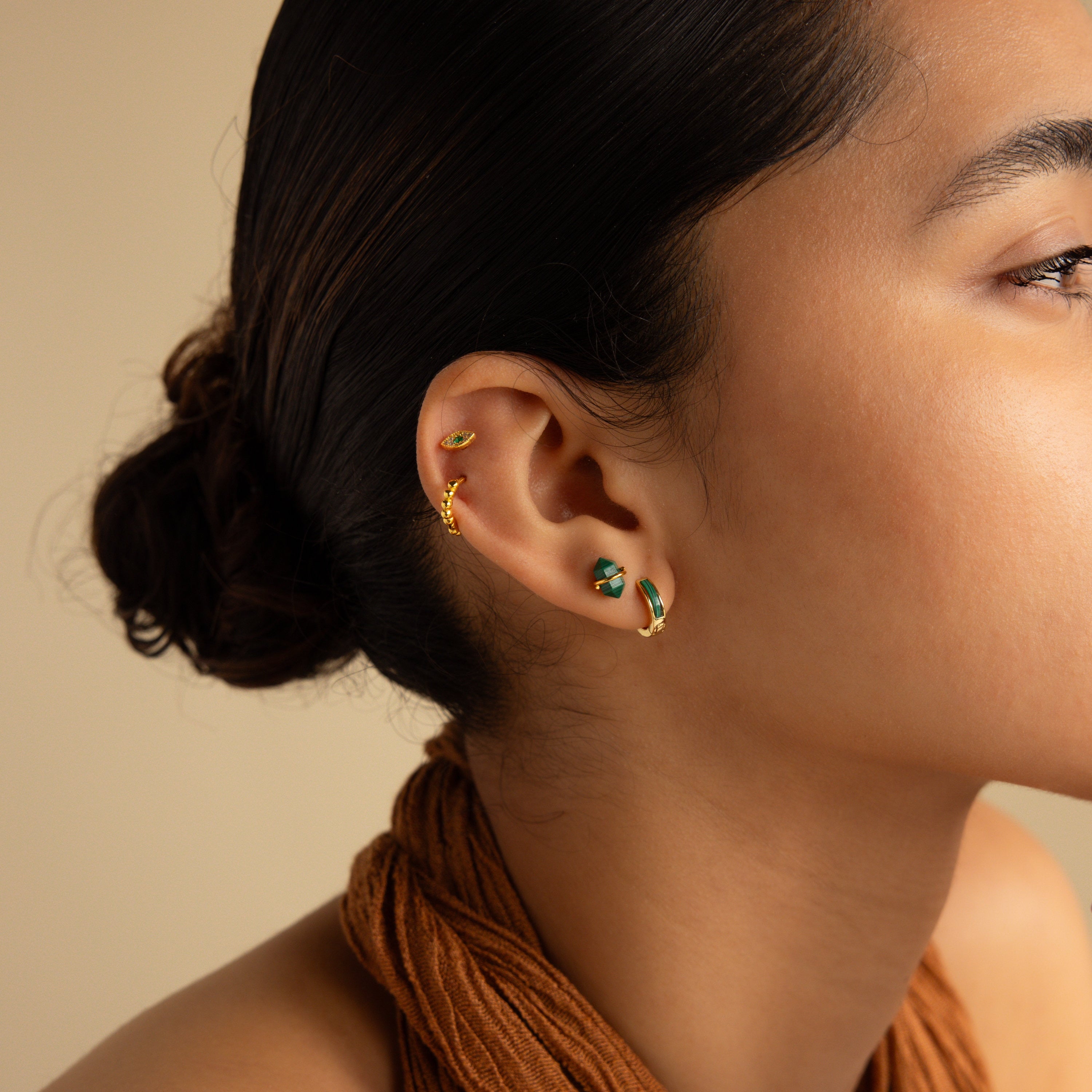 Woman with dark hair in a bun, wearing gold Malachite Inlay Huggies and a brown halter top, shown in side profile to highlight her love for green stone jewelry.
