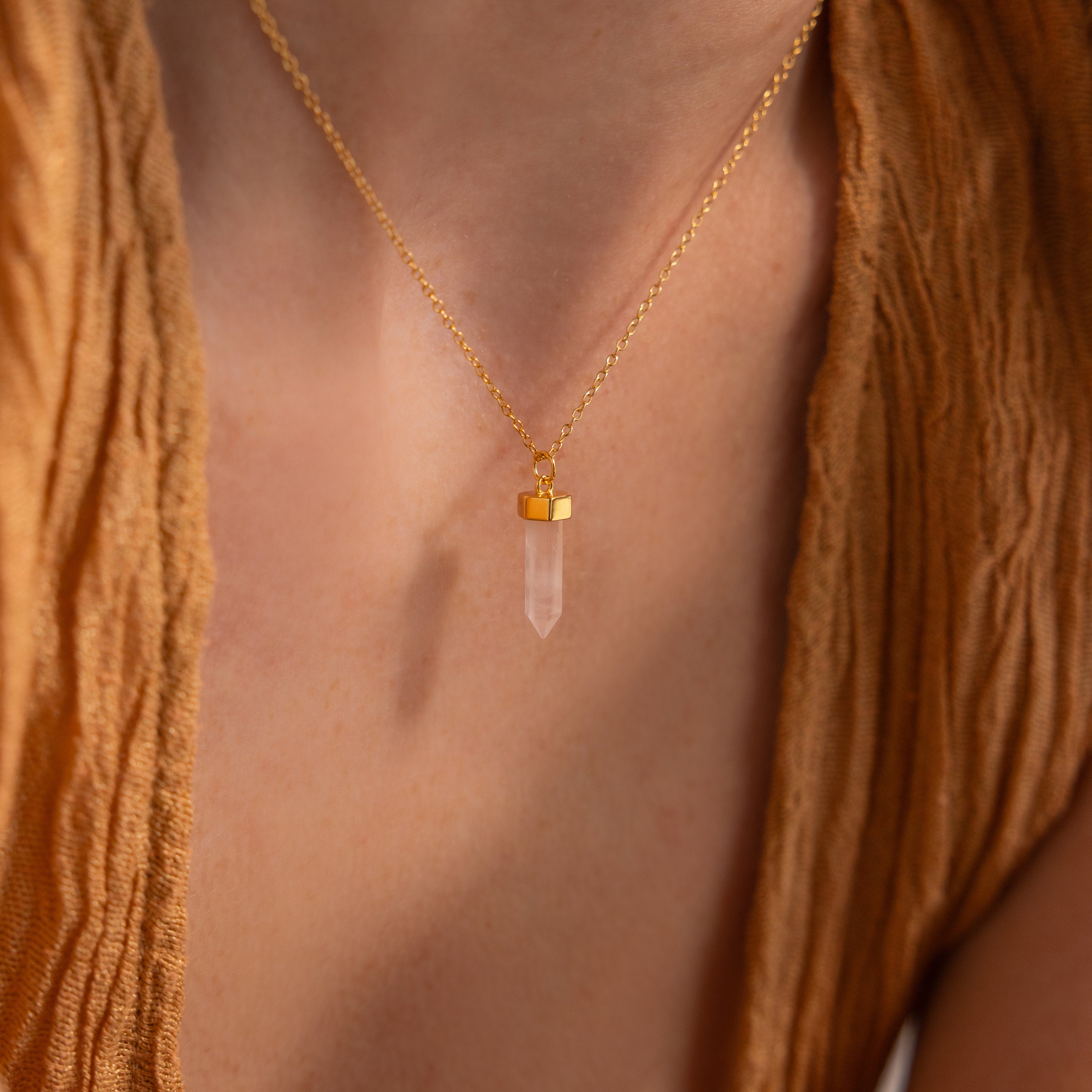 A person wears the Rose Quartz Necklace featuring a gold chain and clear crystal pendant, paired with a textured tan top.