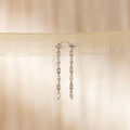 The Dainty Diamond Drop Earrings feature sparkling stones dangling from sheer beige fabric, ideal as wedding or bridesmaid jewelry for an elegant touch against a soft background.