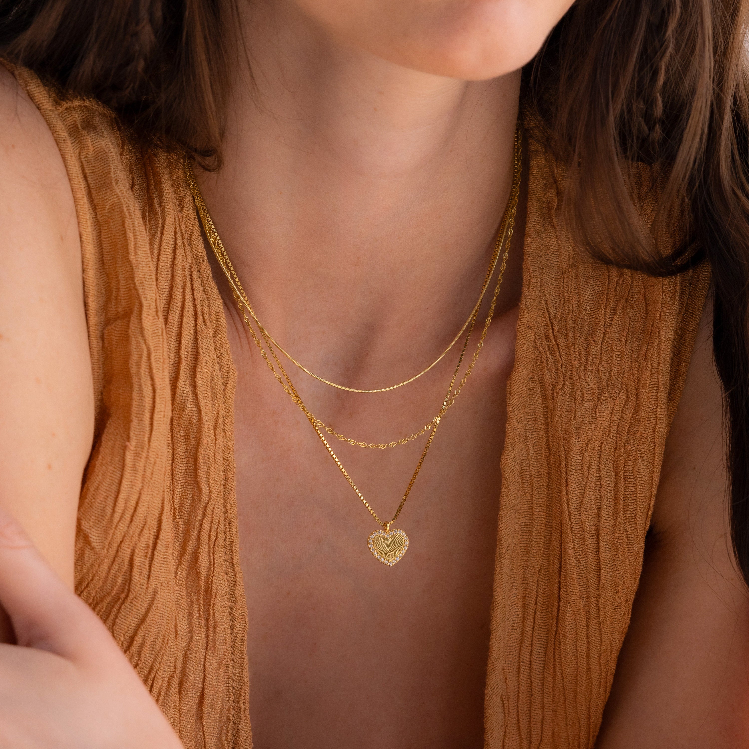 A woman, shown from shoulders to upper chest, wears layered gold necklaces, including the Pave Fingerprint Heart Necklace, styled with a textured tan top.