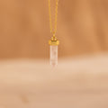 The Rose Quartz Necklace, featuring a gold chain and pointed crystal pendant known as the stone of love, is elegantly displayed against a soft beige background.