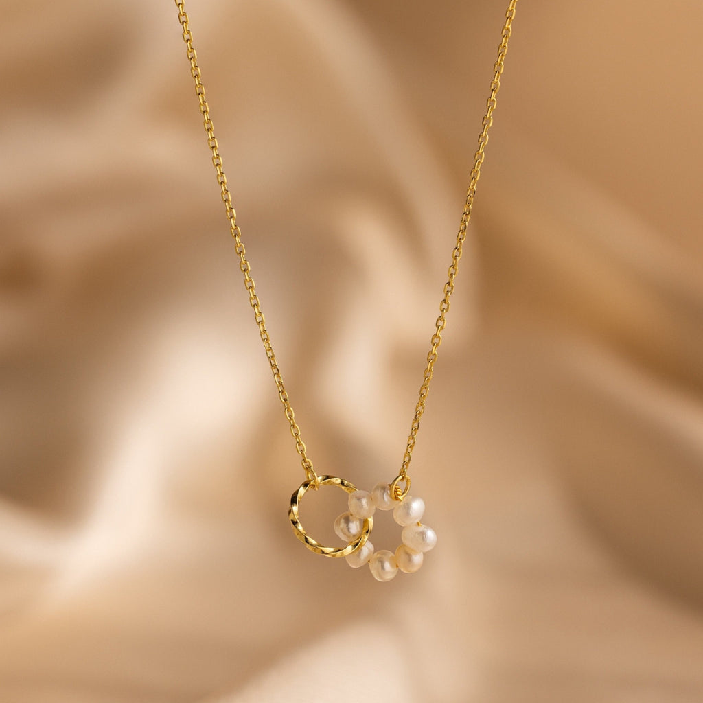 Interlocking Pearl Necklace featuring a gold chain with small bead pearls and a twist ring charm, elegantly displayed on beige satin.