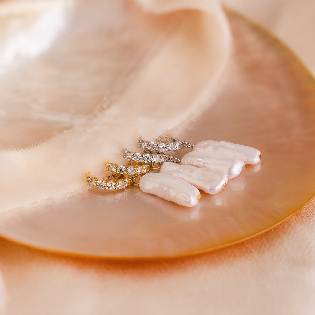 Three pairs of Baroque Pearl Pave Huggies—pearl and crystal drop earrings—are elegantly displayed on a shiny, round shell with soft beige fabric.