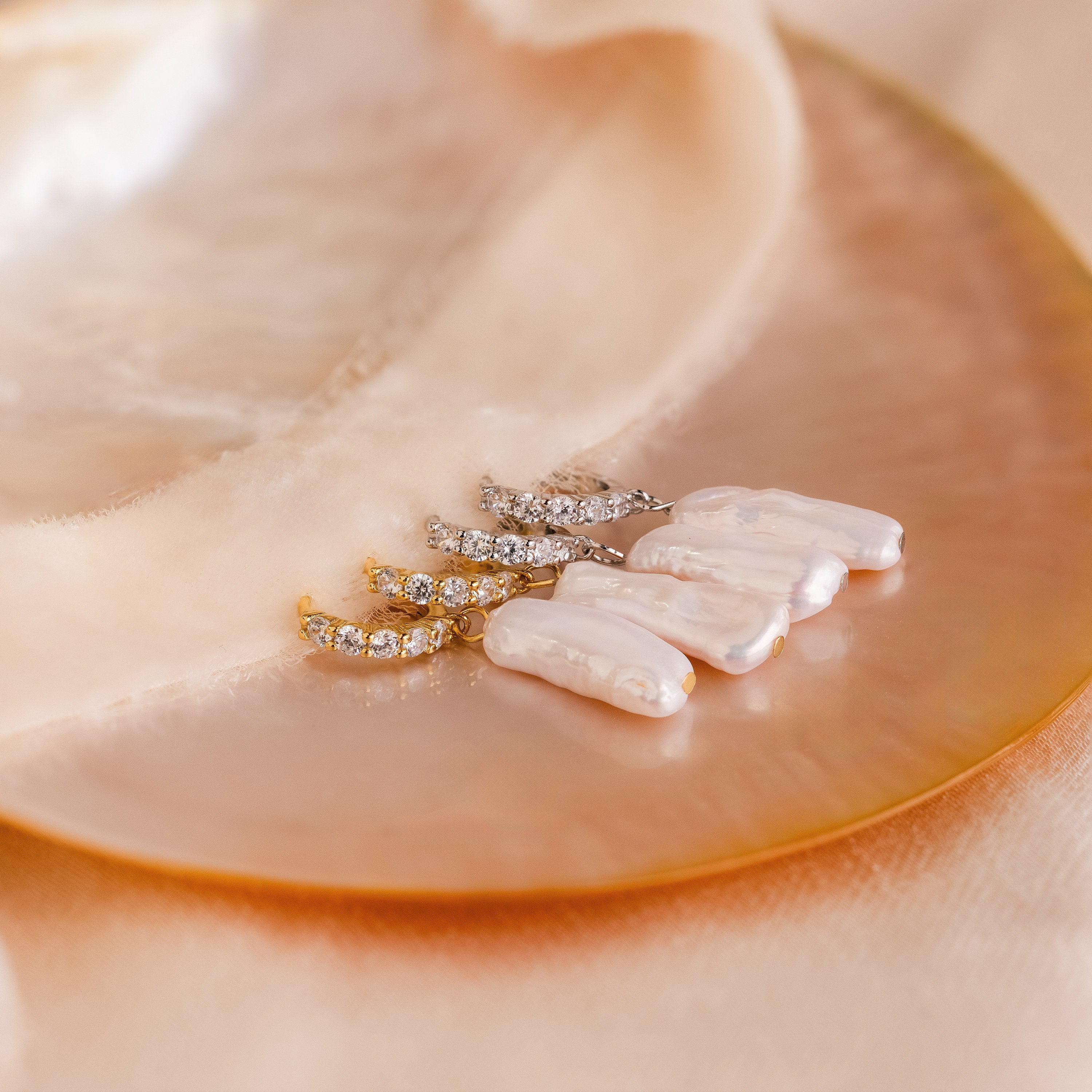 Three pairs of Baroque Pearl Pave Huggies—pearl and crystal drop earrings—are elegantly displayed on a shiny, round shell with soft beige fabric.